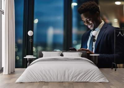 Smiling man in a business suit is looking at his smartphone, standing indoors with the night city lights reflected in the glass window behind him. Wall mural