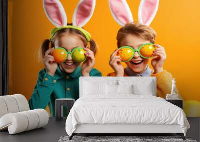 Two joyful children are wearing bunny ears and holding decorated Easter eggs up to their eyes like glasses Wall mural