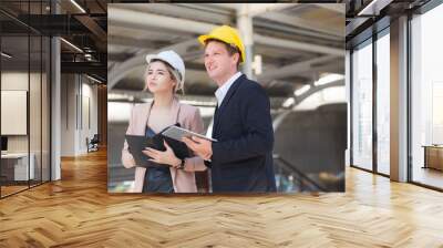 Male and female engineers wear formal suits and hard hats in mobile phones, documents, and tablets. They are looking at and checking the progress of the Sky Train project behind the station. Wall mural