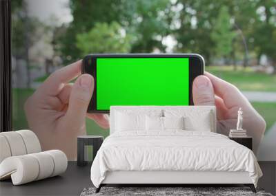 Lublin, Poland - July 2018: Two hands holding a phone with a green screen in a horizontal position in a public park. Close-up shot. Soft focus Wall mural