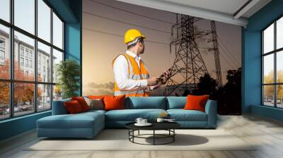 Electrical engineer working. Electrician holding mobile phone (or tablet) and watching on high voltage power of electricity pylon in sunrise Wall mural