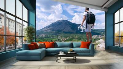 A young man with a backpack holds a drone with remote and looks at the mountain. Wall mural