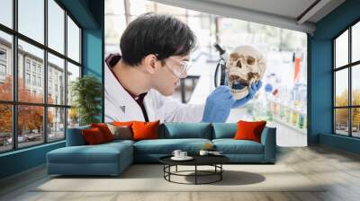 A male scientist with black hair wearing white coat and protective glassware holding a magnifying glass in front of a skull in a white laboratory or hospital. Wall mural