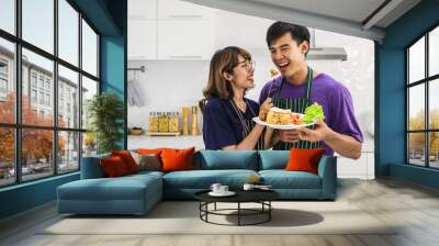 Smiling  happy young Asian couple wearing apron and cooking together and feeding each other in a white kitchen background Wall mural