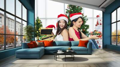 Two young beautiful Asian girls in front of Christmas tree wearing red santa hat opening present for new year or christmas. Wall mural