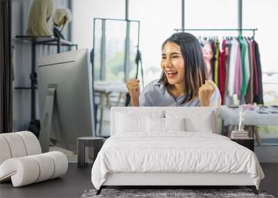 Young Asian woman business owner sitting at a desk in a clothing shop looking at a computer monitor with a happy cheerful expression on her face. Wall mural