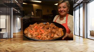 A woman proudly holds a large pan of chicken and carrots in a bustling kitchen Wall mural