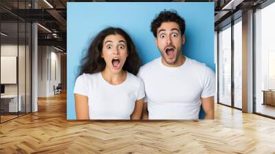 Surprised couple expressing excitement against a blue background in casual attire Wall mural