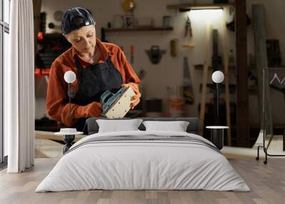 Female carpenter holding sander machine for polishes wooden board while standing in the workshop Wall mural