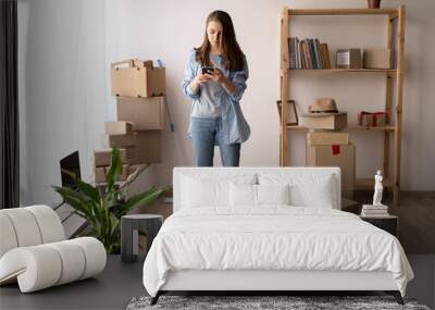 Happy young woman using smartphone in living room at new house with stack of cardboard boxes on moving Wall mural