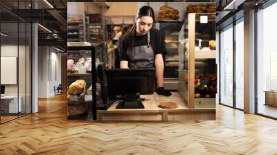 pretty female baker working in bakery shop standing near the showcase of copyspace. Satisfied baker with breads in background. Small business Wall mural