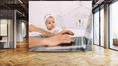 The young dad's hands are on the laptop keyboard, and the little daughter is looking at the screen. They are on the bed. The concept of remote work. Wall mural