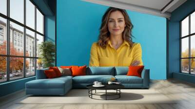 Beautiful, attractive, middle aged woman wearing casual yellow shirt with arms crossed standing isolated on blue background. with generative ai Wall mural