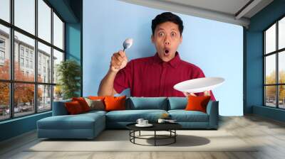 Young Asian man holding to empty eating plate and holding spoon with wow expression Wall mural