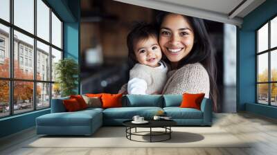 Portrait of a smiling Hispanic mother holding a baby girl in her arms, wearing a beige sweater and white shirt, standing in a home kitchen with a black floor against an empty wall Wall mural