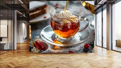 A teacup fills an adorable glass cup with hot tea while cakes with cream clotted around it Wall mural