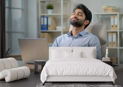 Relaxed young Indian man sitting in office at desk with laptop and documents and resting with closed eyes Wall mural