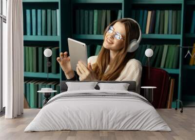 A cheerful female student listens to music and audiobooks in the app, using headphones on her head. Wall mural