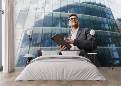 A confident businessman in a suit holds a tablet, standing against a backdrop of modern glass buildings. Wall mural