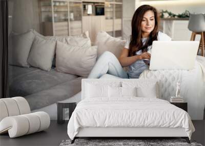 A cozy apartment for a single strong confident woman. A young woman is sitting on a sofa and typing on a computer. A housewife orders clothes home online. A brunette of European appearance. Wall mural