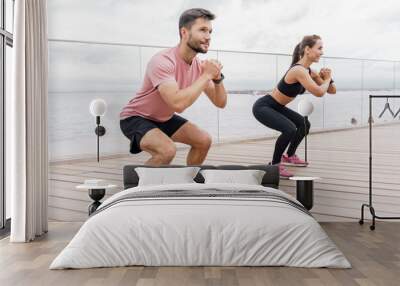 A man and woman perform squats together on a wooden deck by the water, focusing on their workout. Wall mural