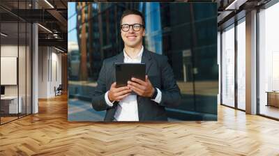 A smiling businessman holding a tablet, standing confidently in front of a modern glass building. Wall mural