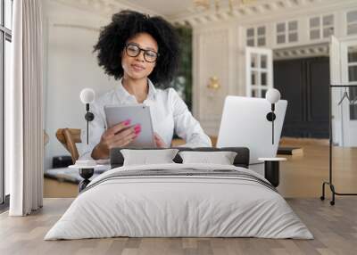 A woman works on the Internet on a computer in an office smart with glasses freelancer lawyer manager assistant uses a tablet stands and looks at the camera smiling. Wall mural