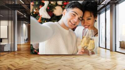 a young couple take a selfie. Christmas tree with a garland and a couple in love are sitting on the couch, celebrating the holiday. new year's card, champagne glasses held in hands. Wall mural