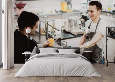 A young man of Caucasian appearance buys coffee with him in a small coffee shop in the city center. A restaurant employee works selling hot drinks to customers. Wall mural