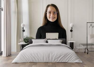 A young woman in a black turtleneck and glasses holds a tablet, smiling warmly in a well-lit, elegant interior. Wall mural