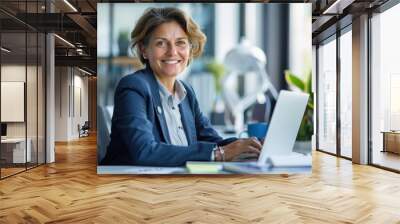 Happy smiling middle aged professional business woman company executive ceo manager wearing blue suit sitting at desk in office working on laptop computer Wall mural