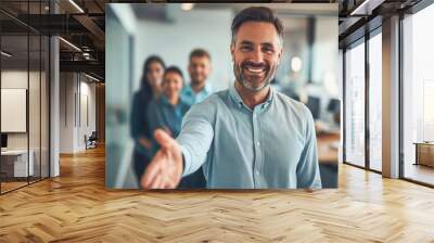 Portrait of businessman welcoming new employee to his business team and company, giving hand forward in modern office with colleagues in the background Wall mural