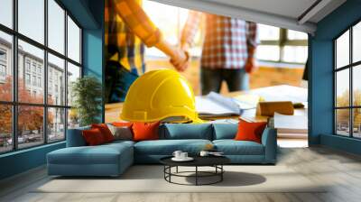 Yellow safety helmet on workplace desk with construction worker team hands shaking greeting start up plan new project contract in office center at construction site, partnership and contractor concept Wall mural