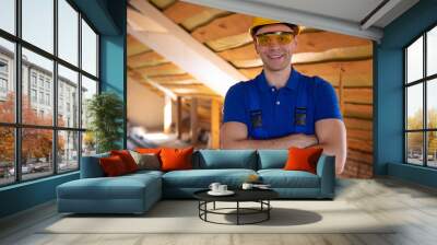 A smiling construction worker in a yellow hard hat and safety glasses crosses his arms in an unfinished attic. The confident expression suggests satisfaction with the progress of the renovation. Wall mural