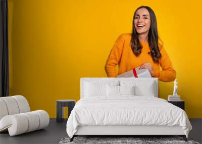 Attractive excited smiling young woman holds beautiful gift box in hands and looks on the camera isolated on yellow background Wall mural