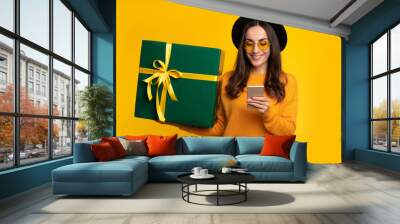 Attractive excited smiling young woman holds beautiful gift box in hands and looks on the camera isolated on yellow background Wall mural