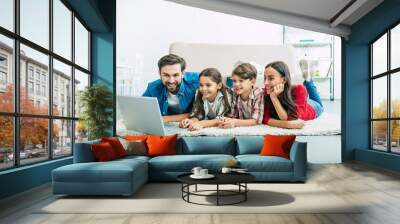 Beautiful modern young family lying on the floor at home and doing something in laptop Wall mural