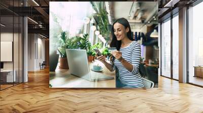 Beautiful smiling young woman using laptop and doing shopping online with help of her smartphone and card Wall mural