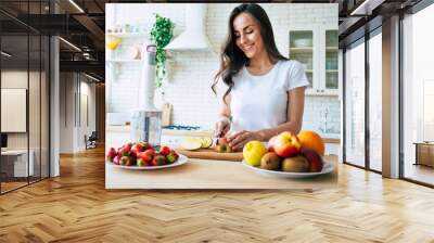 Beautiful woman making fruits smoothies with blender. Healthy eating lifestyle concept portrait of beautiful young woman preparing drink with bananas, strawberry and kiwi at home in kitchen. Wall mural