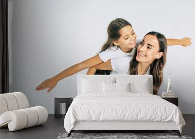 Beautiful young brunette mother holds on back her cute little daughter in white T-shirts isolated in studio Wall mural