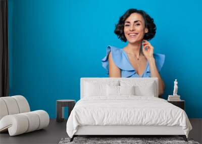 Close-up photo of a charming healthy woman in a blue dress, who is looking in the camera and smiling, while standing and posing isolated on blue background Wall mural