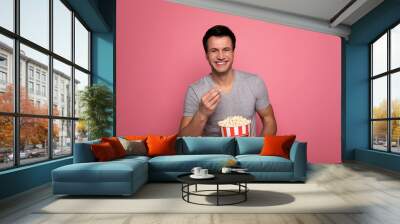 Comedy lover. Close-up photo of a handsome young man, who is looking in the camera and laughing, while eating popcorn. Wall mural