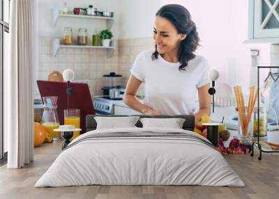 Cute beautiful and happy young brunette woman in the kitchen at home is preparing fruit vegan salad or a healthy smoothie and having fun Wall mural