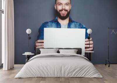 Everything is for you. Handsome young smiling man in a denim shirt shows a white sheet of paper in the camera on a gray background. Area for advertising. Wall mural