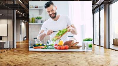 Handsome happy bearded man is preparing wonderful fresh vegan salad in the kitchen at home Wall mural