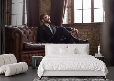 Handsome stylish confident bearded businessman in a smart suit and glasses sitting on a sofa in the office. Wall mural