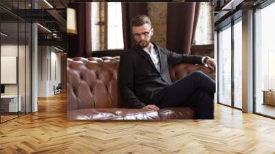 Handsome stylish confident bearded businessman in a smart suit and glasses sitting on a sofa in the office. Wall mural