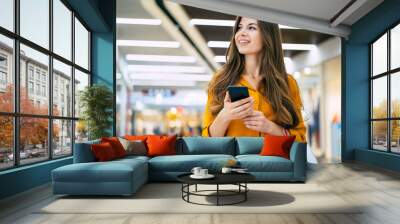 Happy beautiful young stylish woman with shopping bags is using smart phone while walking in the mall on black friday Wall mural