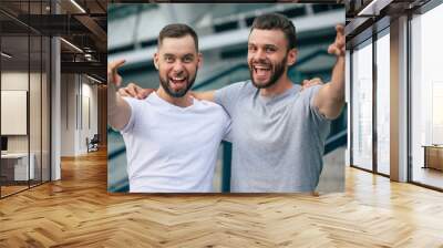 Happy guys pointing on camera. Two young bearded handsome friends in colorful t-shirts are posing together on the city background. Wall mural