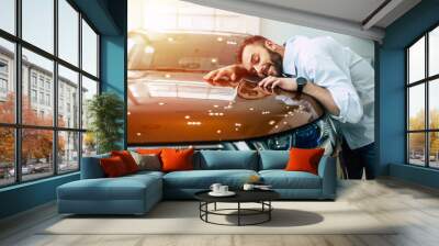 Happy handsome bearded man buying a car in dealership, guy hugging hood of new car Wall mural